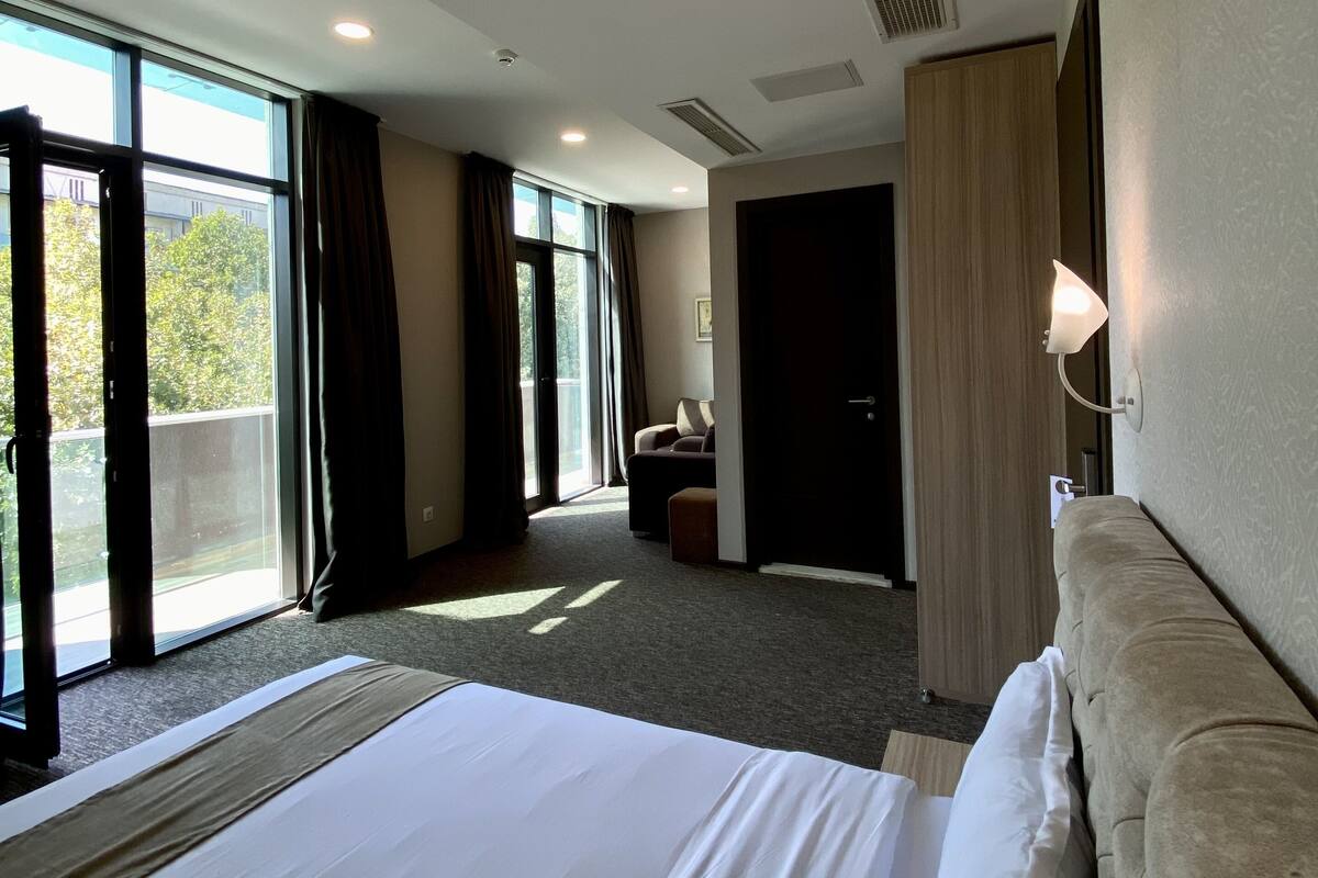 A hotel room with a large bed, a sofa, a desk, and a view of the outdoors through the windows.