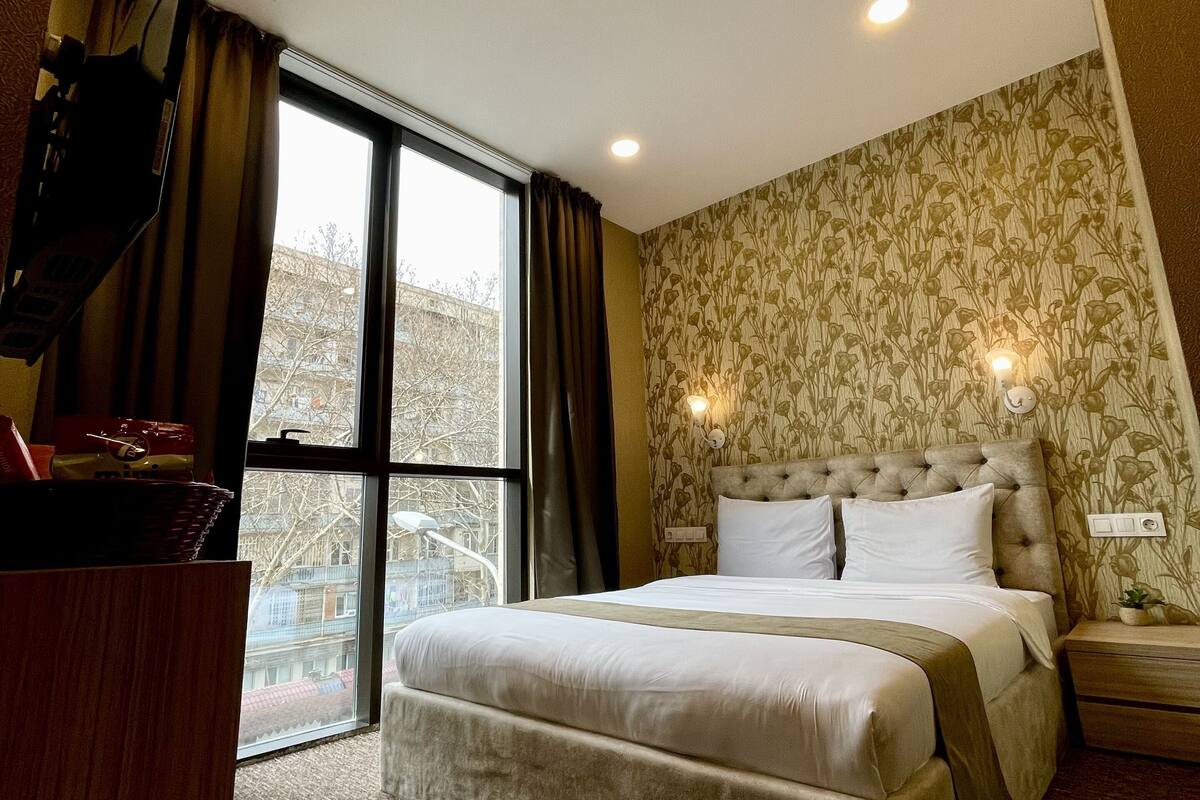 A hotel room with a large bed, a television, a window with curtains, and a patterned wall.