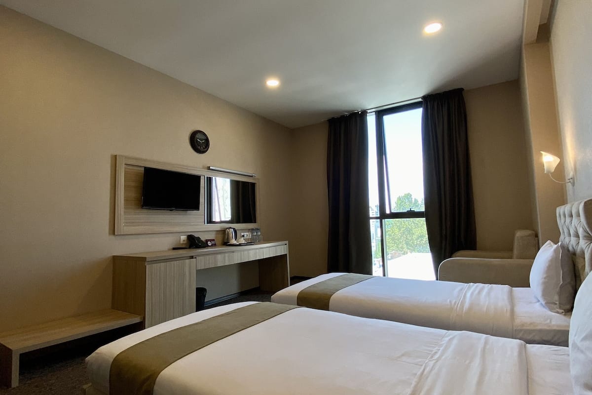 A hotel room with two beds, a desk with a flat-screen TV, a clock, and a window with curtains.