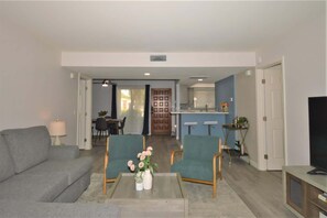 Ein Wohnzimmer mit einer grauen Couch, zwei grünen Sesseln, einem Couchtisch mit Blumen und einem Fernseher auf einem Holzboden.