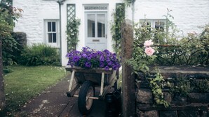 A white building with a sign reading "WEATHERCOCK" and a wheelbarrow filled with purple flowers in front. - Luxurious Cottage with Hot Tub. 19 miles from Windermere. (Sedbergh)