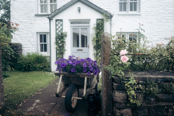 A white building with a sign reading "WEATHERCOCK" and a wheelbarrow filled with purple flowers in front. - Superb Dales Cottage with enclosed garden. 19 miles from Windermere. (Sedbergh)
