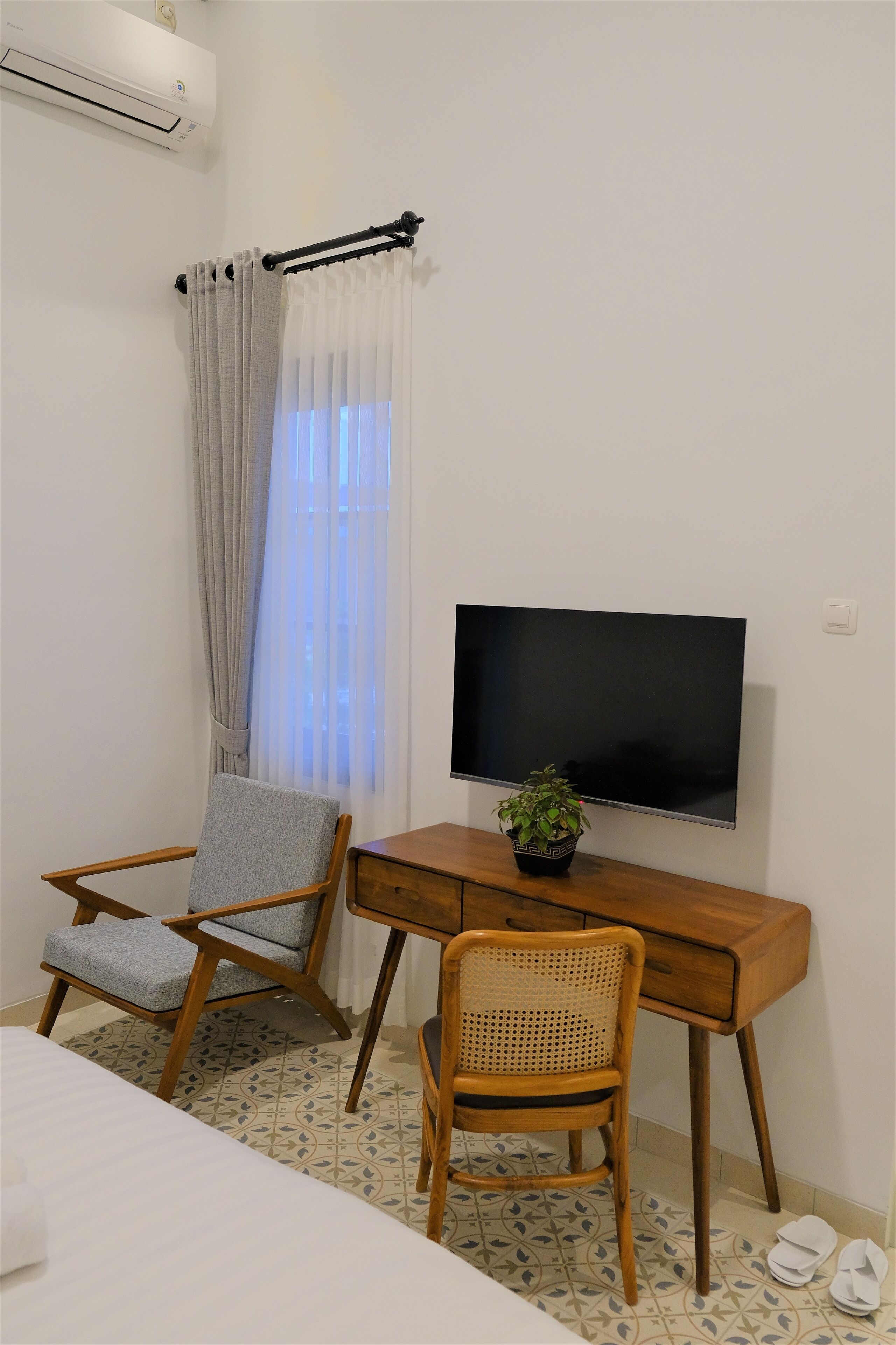 Une chambre d’hôtel avec un lit, deux chaises, un bureau avec une télévision, une plante en pot et une fenêtre avec des rideaux.