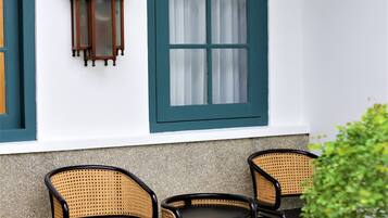 Una habitación con pared blanca, ventana con detalles verdes, dos sillas de mimbre y una pequeña mesa.