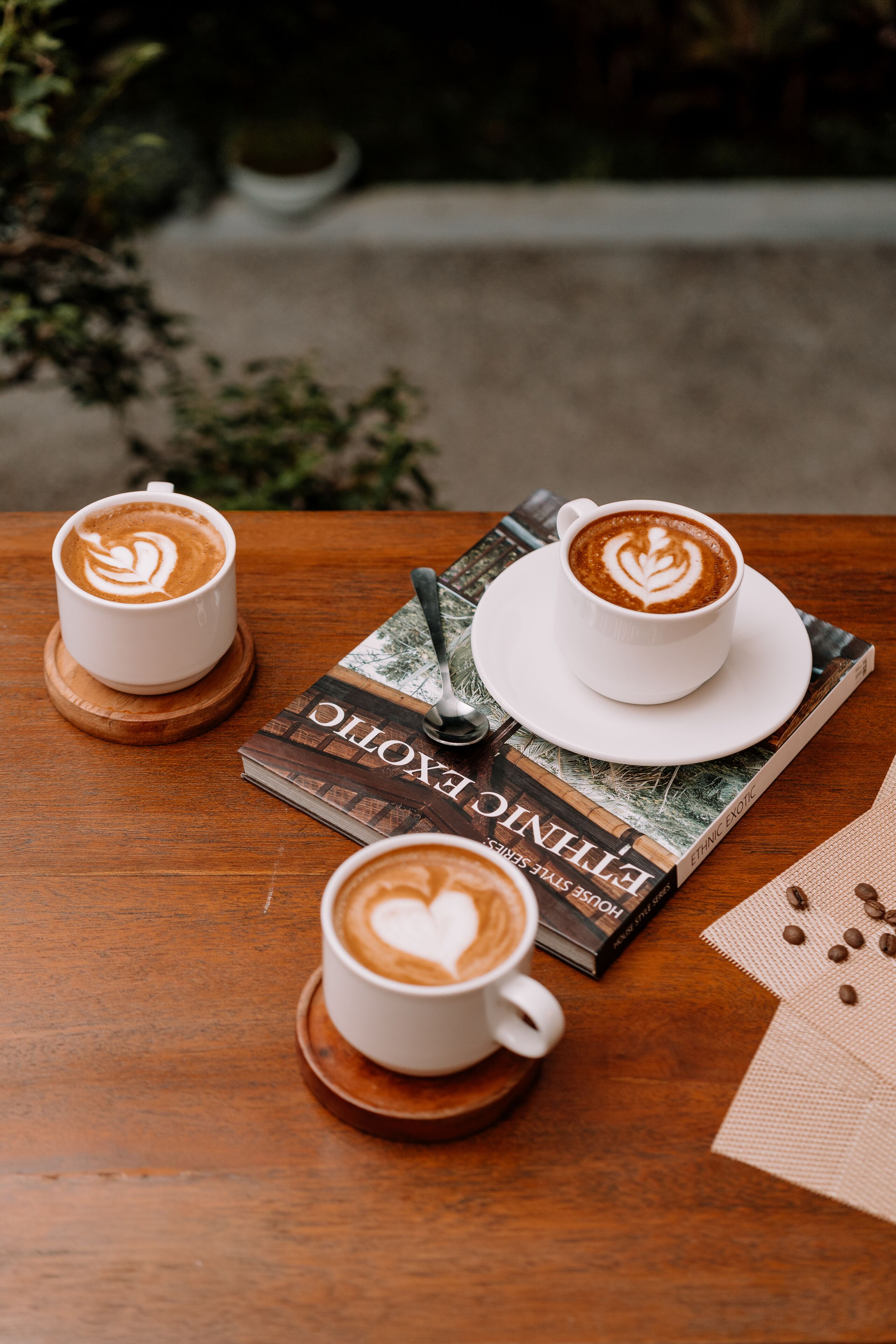 Two cups of coffee with latte art on a wooden table, a spoon, a book titled 'Ethical House', and a napkin with coffee beans.