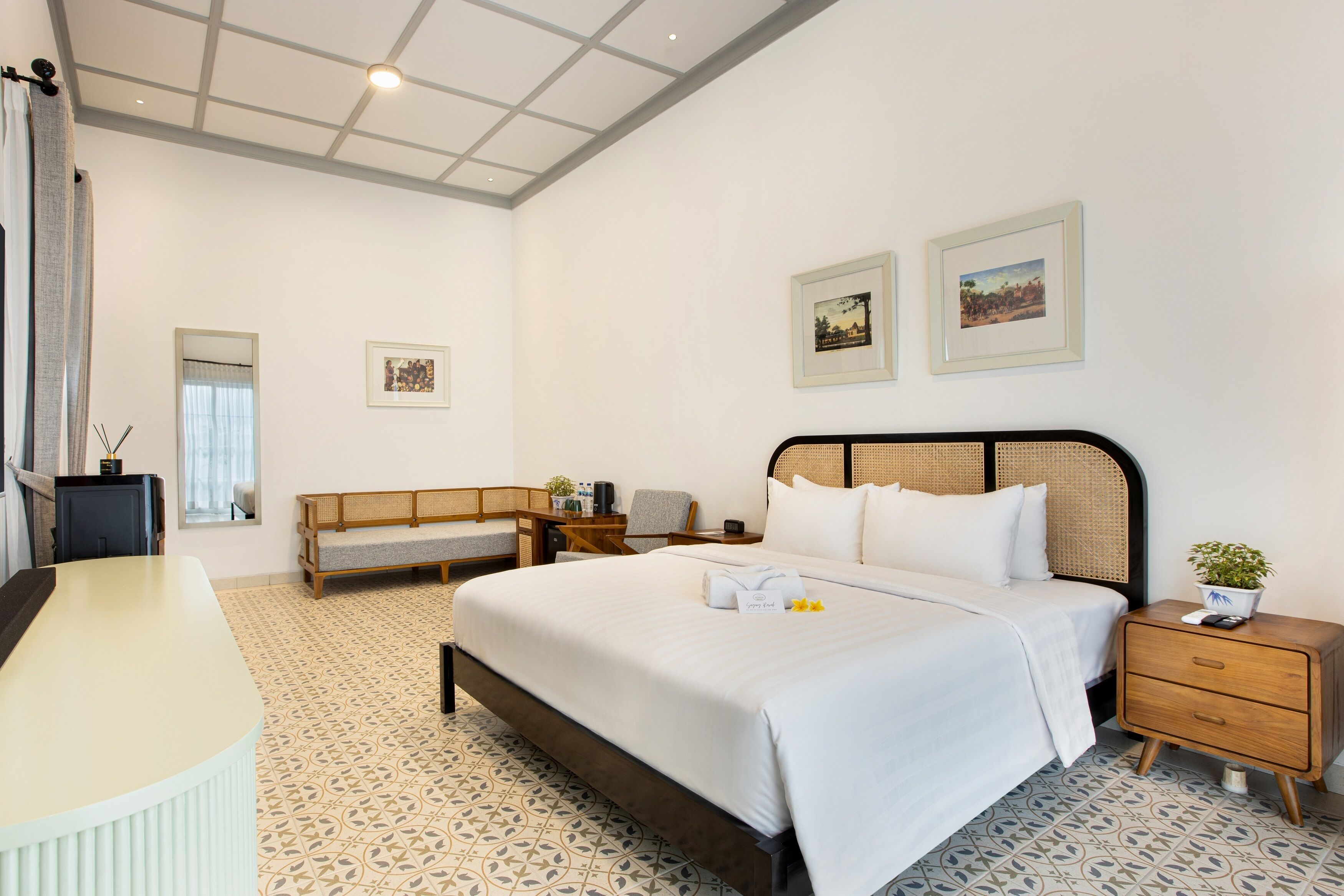 A hotel room with a large bed, a desk, a chair, a small table, and two framed pictures on the wall.