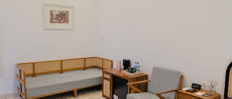 A room with a grey sofa, a wooden desk, a chair, a bedside table with a lamp, and a framed picture on the wall.