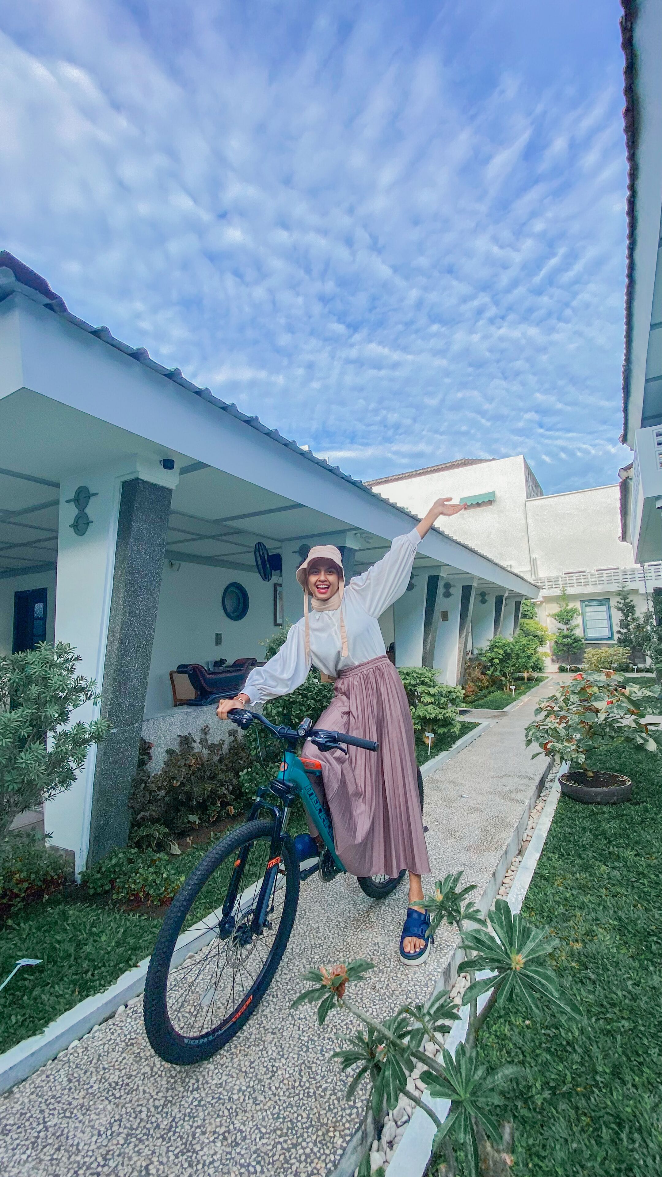A person on a bicycle in a courtyard with a white building, greenery, and a clear sky.