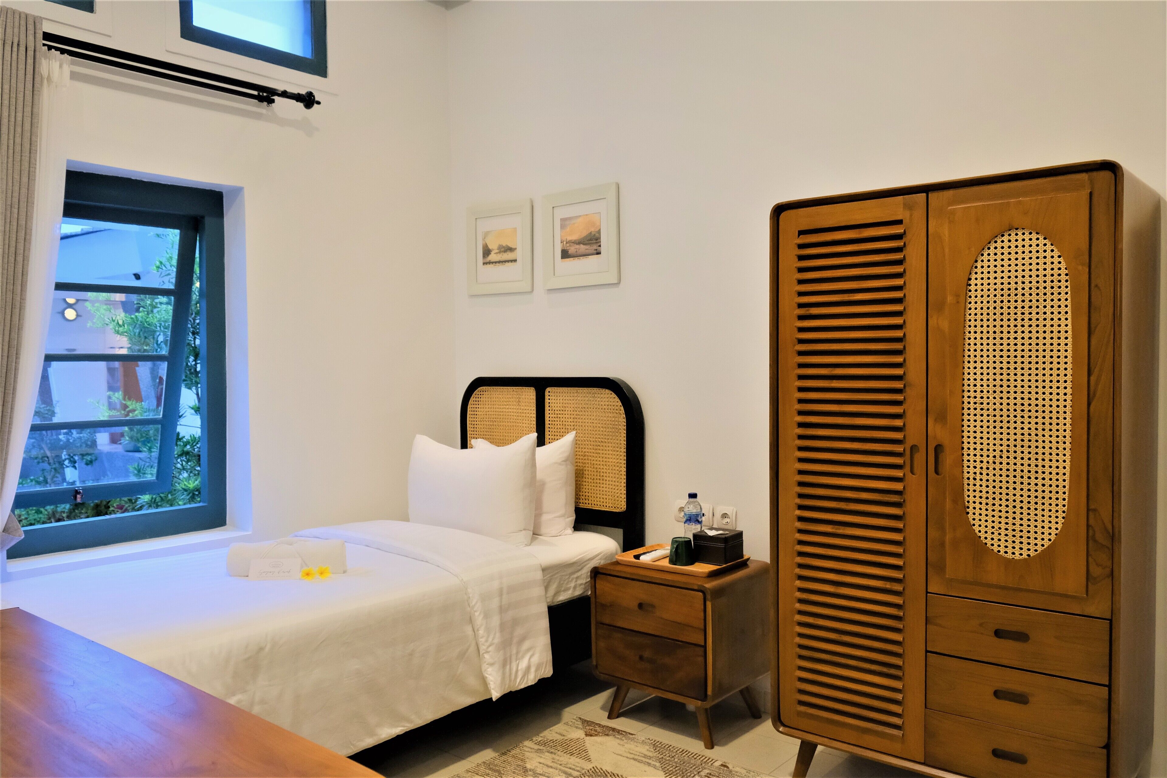 A hotel room with a bed, a wooden wardrobe, a nightstand, and a window with a view of greenery.