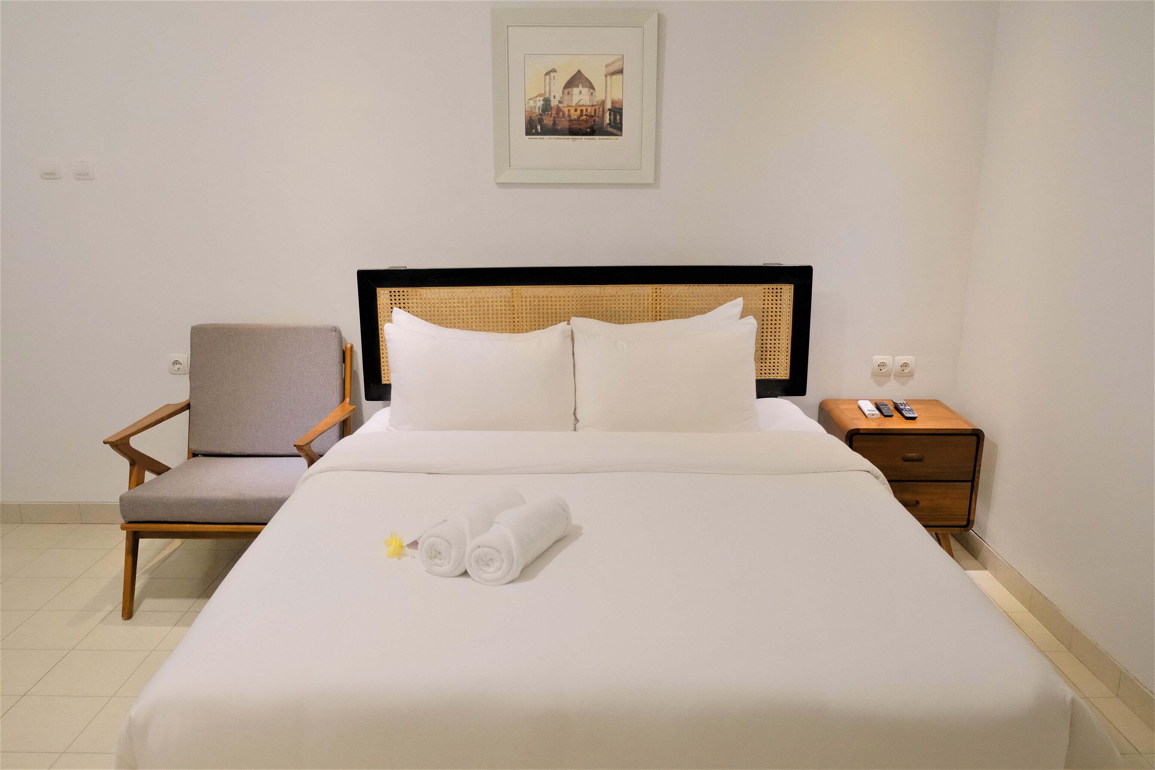 A neatly made bed with white linens, a wooden nightstand with a remote control, a grey upholstered chair, and a framed picture on the wall.