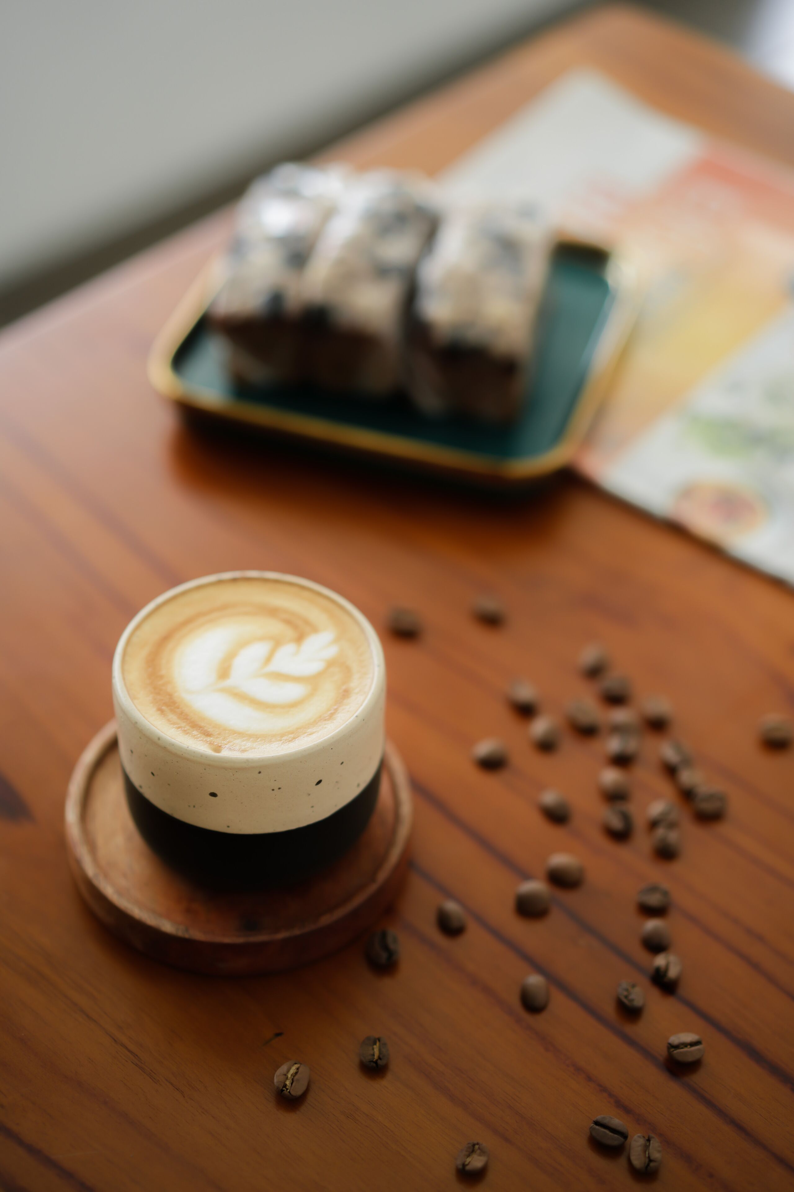 A cup of coffee with latte art on a wooden table, scattered coffee beans, and a plate of pastries in the background.