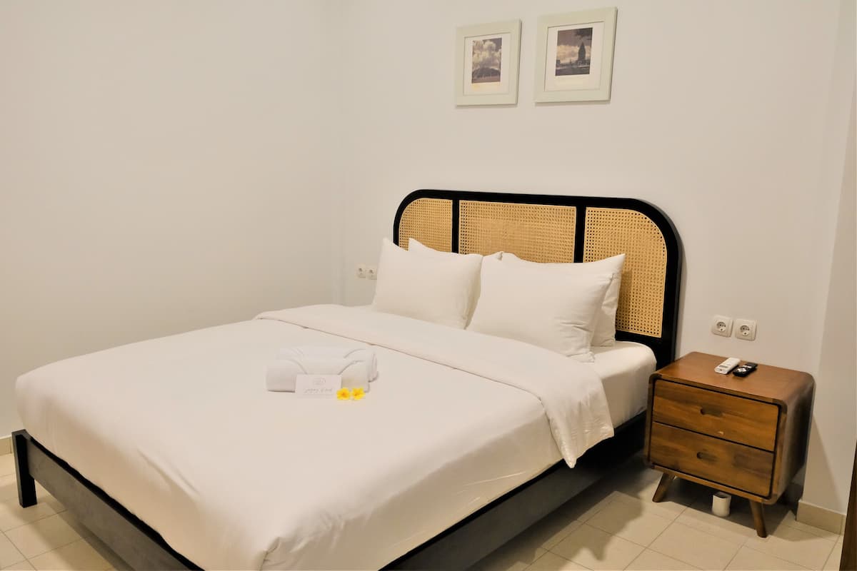 Een net opgemaakt bed met witte lakens en kussens, een nachtkastje met een afstandsbediening, en twee ingelijste foto's aan de muur.