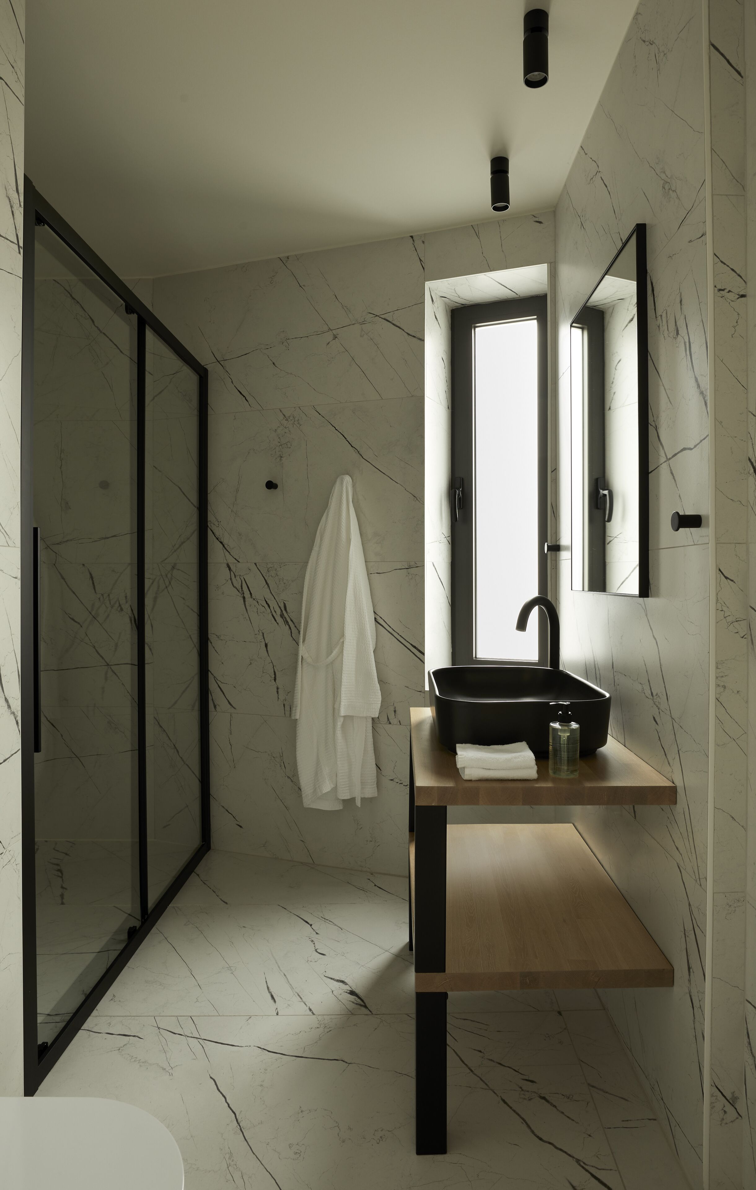 A modern bathroom with a marble floor, a wooden vanity, a black sink, a mirror, and a towel hanging on the wall.