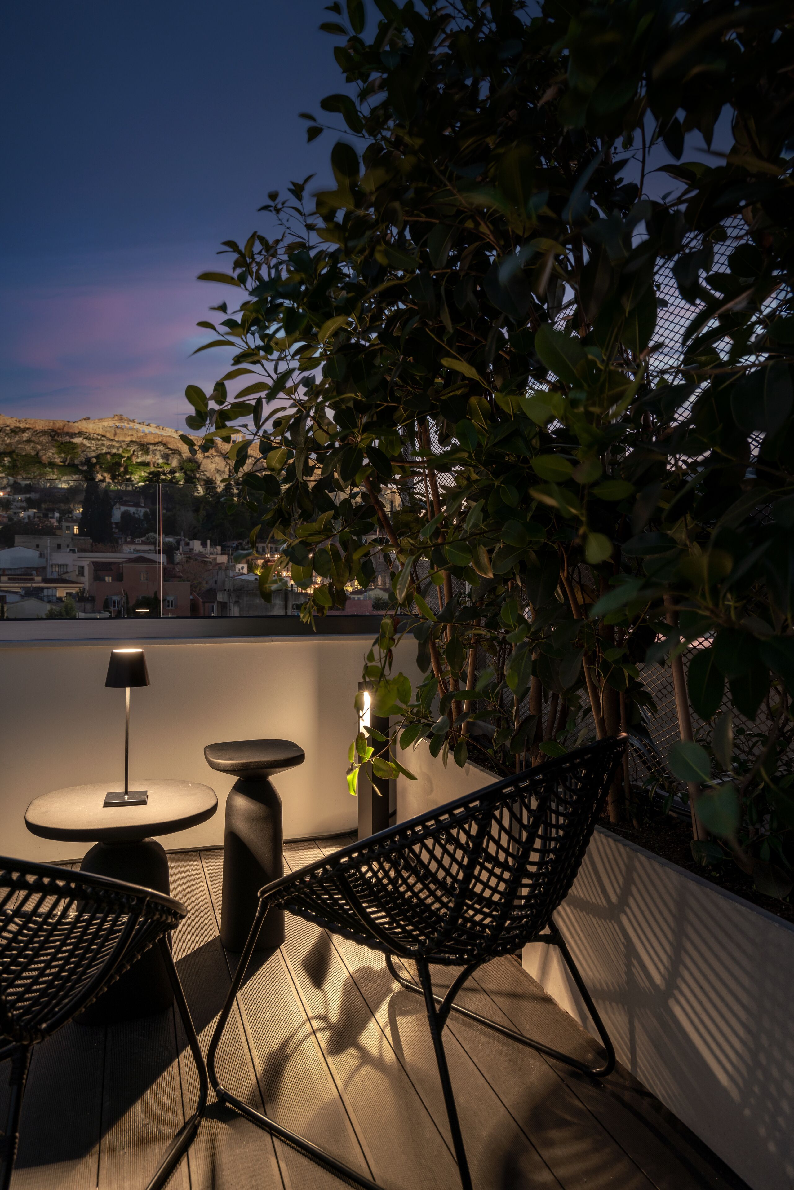 En tagterrasse med et bord, stole, en lampe og udsigt over bybilledet i skumringen.