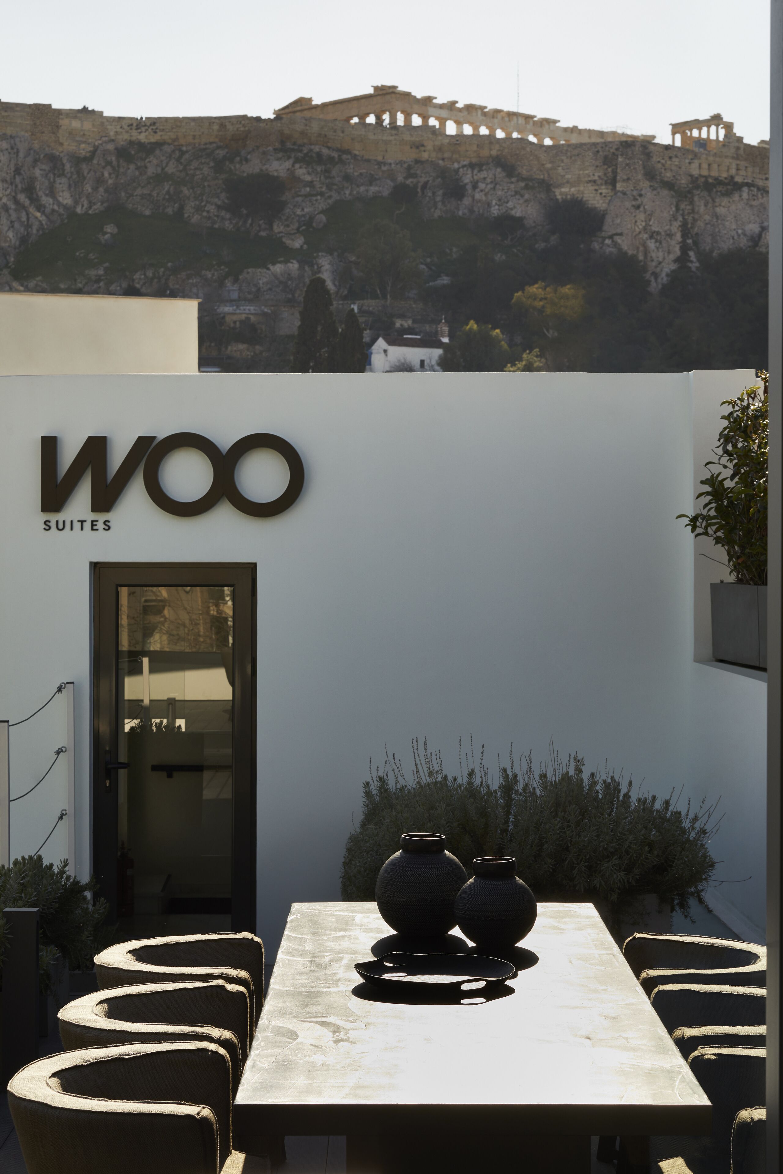 Un espace extérieur moderne aménagé avec une table et des chaises, une enseigne indiquant « Woo Suites », et en arrière-plan, une vue sur d’anciennes ruines.