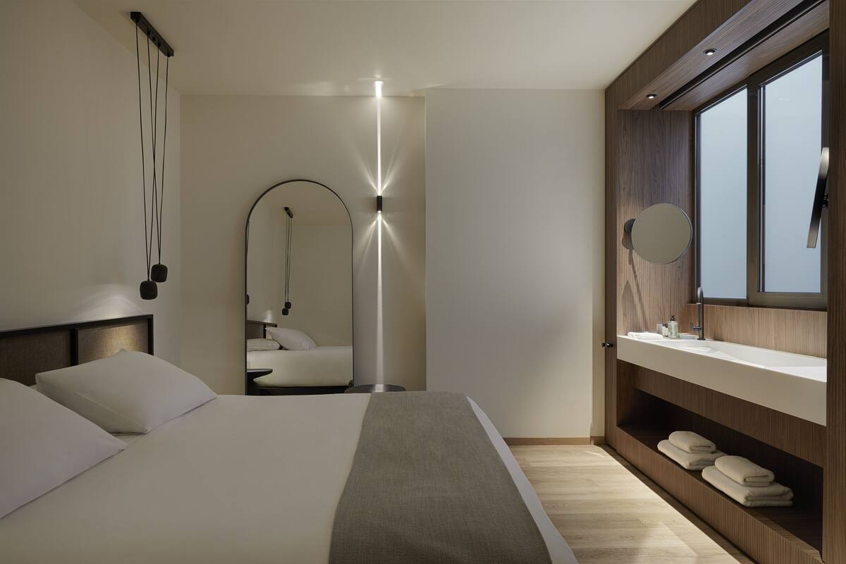 A modern hotel room with a large bed, a bedside table, a mirror, a built-in vanity with a sink, and a large window.