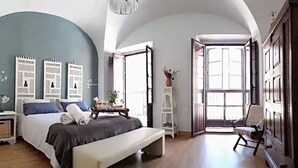 A bedroom with a large bed, a bench, a chair, a white shelf, and a large window.