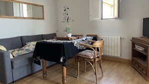 A living room with a sofa, a wooden dining table with a black tablecloth, a chair, a potted plant, and a television on a wooden stand.