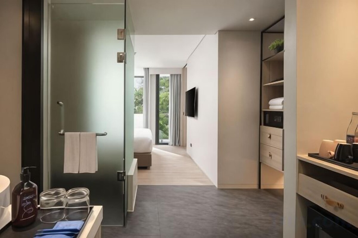 Une chambre d’hôtel moderne dotée d’une porte en verre donnant sur la salle de bain, d’un lit, d’un téléviseur fixé au mur et d’un petit coin cuisine.