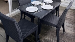 Un área de comedor al aire libre con una mesa dispuesta para cuatro personas, rodeada de sillas de mimbre, sobre una terraza de madera.