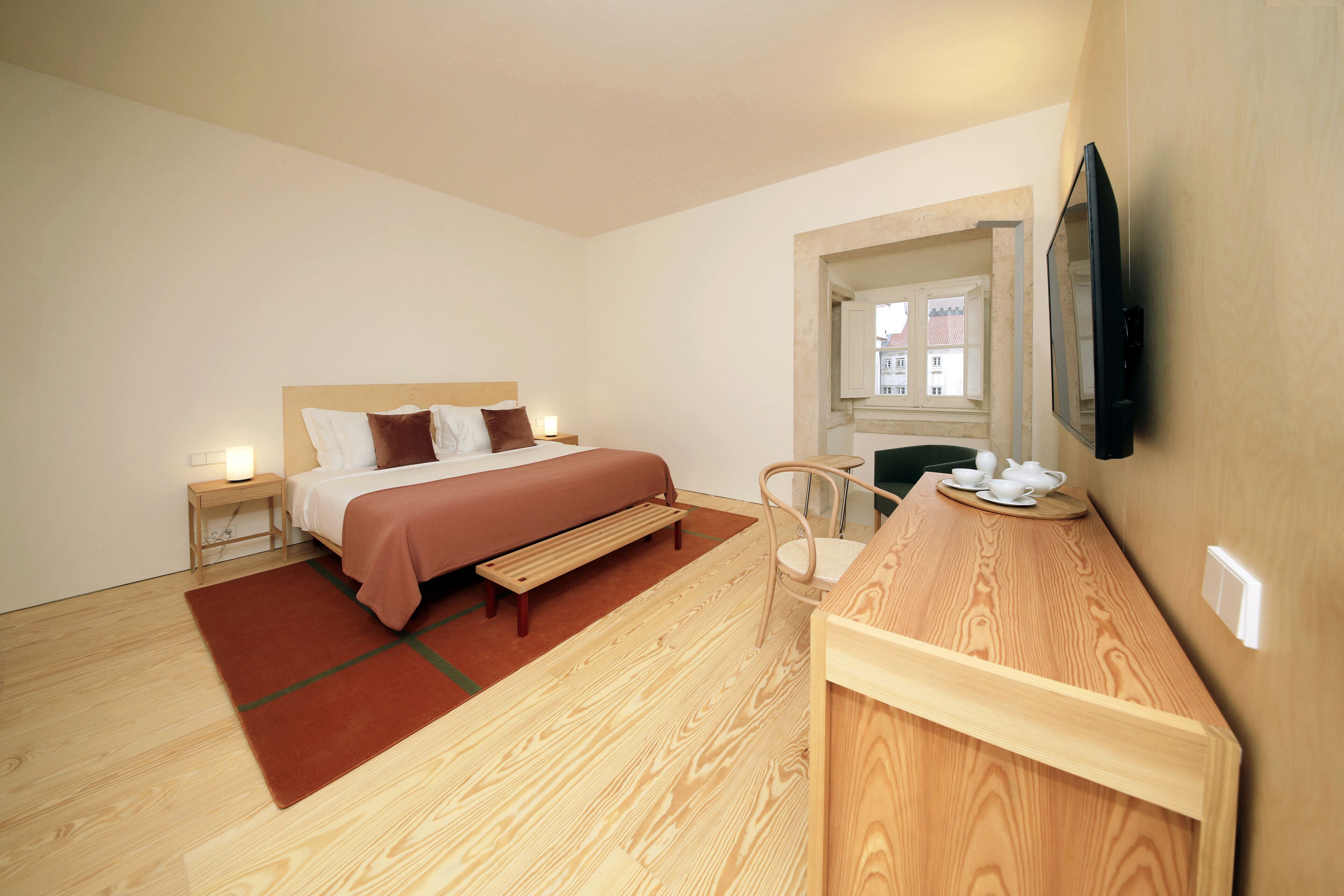 A modern hotel room with a bed, bedside table, chair, and a wooden desk with a mirror.
