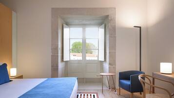 Une chambre à coucher comprenant un lit, un bureau, une chaise, une petite table et une fenêtre donnant sur les bâtiments environnants.