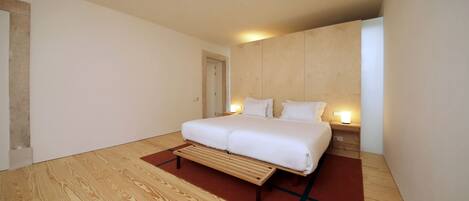 A modern bedroom with a wooden floor, a bed with white bedding, and a nightstand with a lamp.
