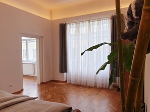 Een kamer met een visgraat parketvloer, een bed met een witte hoes, een grote groene plant en een raam met transparante gordijnen.