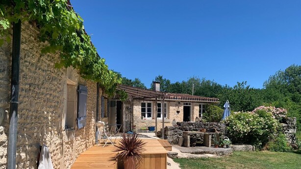 Un bâtiment en pierre doté d’une terrasse en bois, d’un mur en pierre et d’un jardin avec des plantes et un banc.
