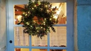 Une porte blanche ornée d’une couronne de Noël et de guirlandes lumineuses, une fenêtre avec des rideaux en dentelle et un sol carrelé.