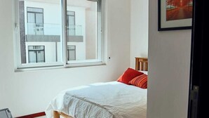 Een slaapkamer met een groot raam, een bed met witte beddengoed en rode kussens, en een ingelijst beeld aan de muur.