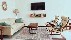Een woonkamer met een bank, twee fauteuils, een salontafel, een boekenkast en een flatscreen televisie aan de muur.