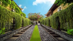 Uma trilha de pedras leva a um edifício tradicional com telhado de azulejos, rodeado por vegetação exuberante e um céu azul límpido.
