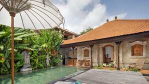 Área de piscina com deck de madeira, estátua de pedra e vegetação exuberante.