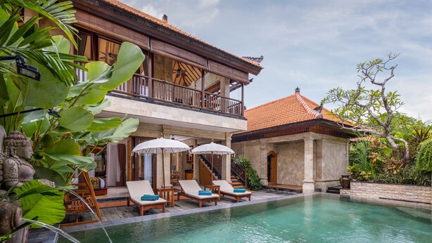 Uma área de piscina com espreguiçadeiras e guarda-sóis, uma escadaria que leva a uma varanda e um edifício com telhado em azulejos.