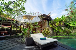 A lounge chair by a pool with a canopy umbrella, surrounded by lush greenery and a traditional building in the background.