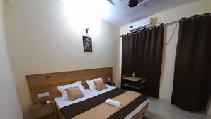A neatly arranged bedroom with a wooden headboard, a bed with white and brown bedding, a bedside table with a lamp, and a small table with a chair. - Hotel Krishna Rishikesh (Narendranagar)