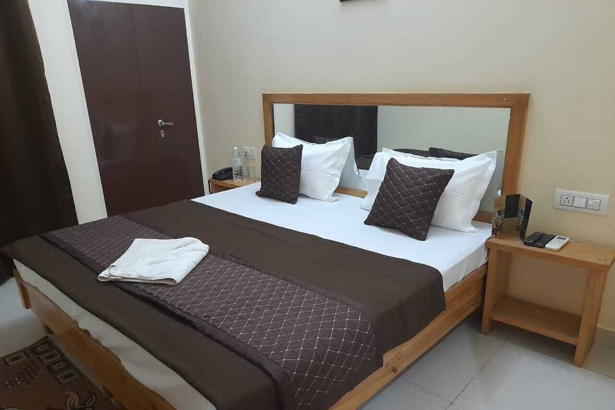 A bed with a wooden frame, white and brown bedding, a mirror above the bed, a wooden nightstand with a phone, and a door in the background.