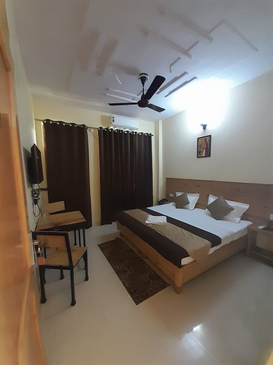 A hotel room with a bed, a desk, a chair, a ceiling fan, and a wall-mounted light fixture.