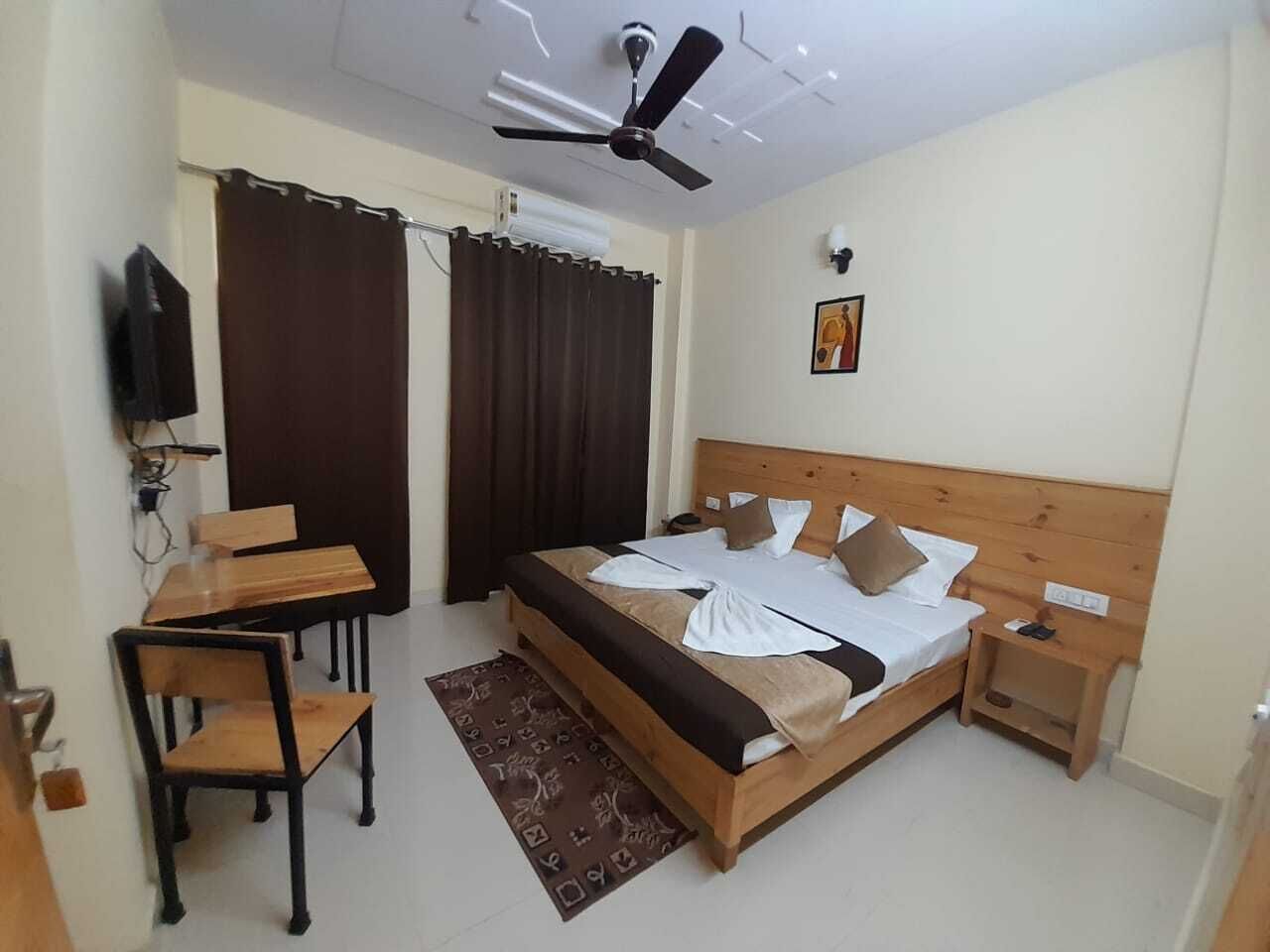 A bedroom with a bed, bedside tables, a desk, chairs, a ceiling fan, curtains, and a wall-mounted TV.