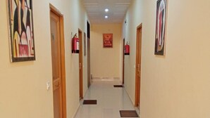 A corridor with doors on both sides, fire extinguishers mounted on the walls, and a central corridor leading to a room with a red and gold framed picture on the wall. - Hotel Krishna Rishikesh (Narendranagar)