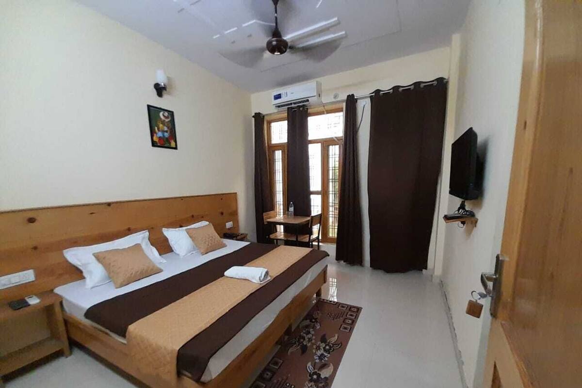 A hotel room with a wooden bed, a ceiling fan, an air conditioning unit, a television, and a window with curtains.