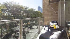 Un balcon doté d’une balustrade en verre, une table avec des aliments pour le petit-déjeuner, et une vue sur des arbres et un bâtiment en arrière-plan.