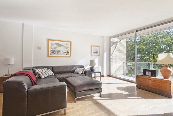 A modern living room with a sectional sofa, a wooden side table, and a large window with a view of trees. - Beautiful apartment near the beach in Barcelona (Barcelona)
