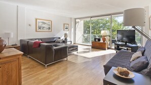Un salon moderne avec un canapé, une table basse et un téléviseur. Une grande fenêtre offre une vue sur les arbres à l’extérieur.