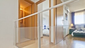 Une chambre d’hôtel moderne avec une cloison en verre, une porte en bois, un lit recouvert d’une courtepointe à pois, une table de chevet et une fenêtre avec des rideaux.