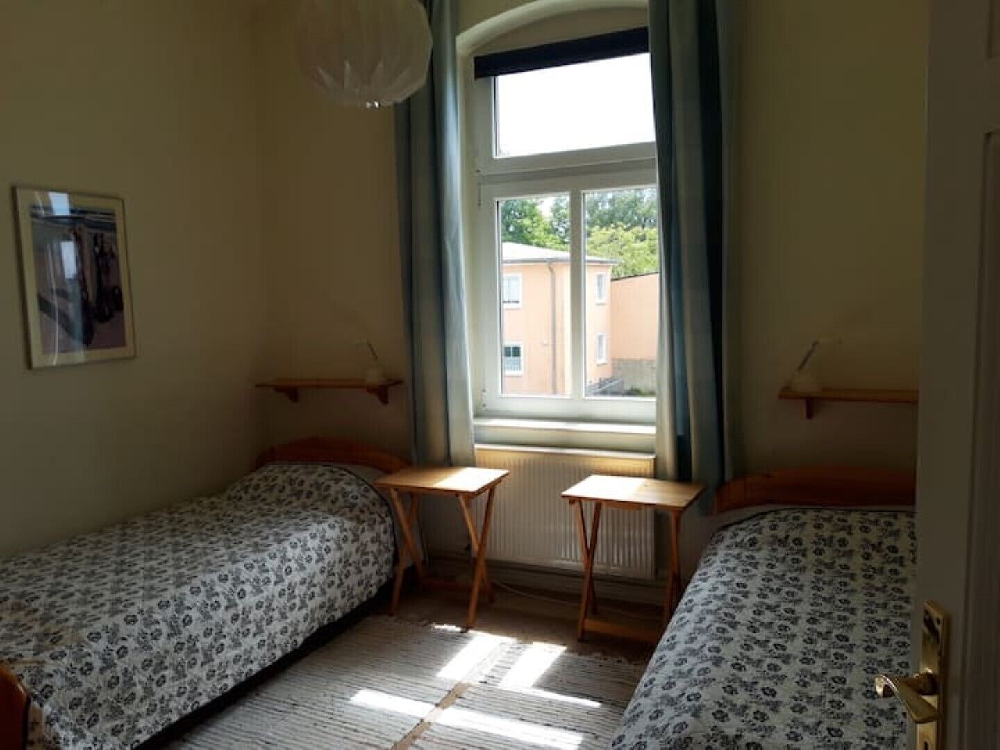 Une petite pièce avec deux lits simples, une petite table, un radiateur et une fenêtre avec des rideaux.
