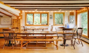 Un coin repas en bois, avec une grande table et des chaises, de grandes fenêtres donnant sur un paysage verdoyant et un plafond rustique aux poutres apparentes.