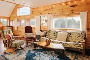 L’intérieur chaleureux d’un chalet, avec un décor en bois, un canapé, un fauteuil, une table et une fenêtre donnant sur une forêt.
