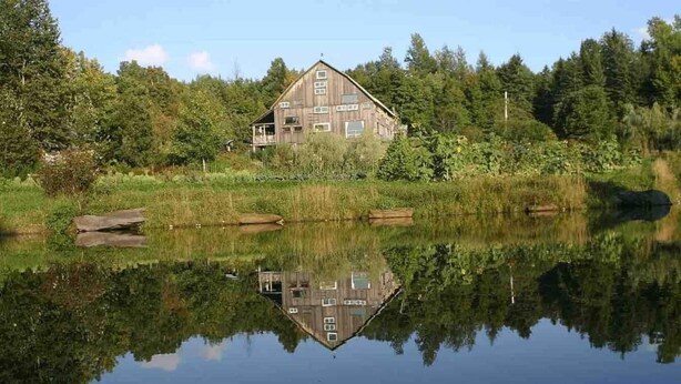A rustic wooden house with a large porch, surrounded by dense forest and a calm lake reflecting its structure. - 100+ Acre Farm Retreat for Groups, Yoga, Weddings (Greensboro Bend)
